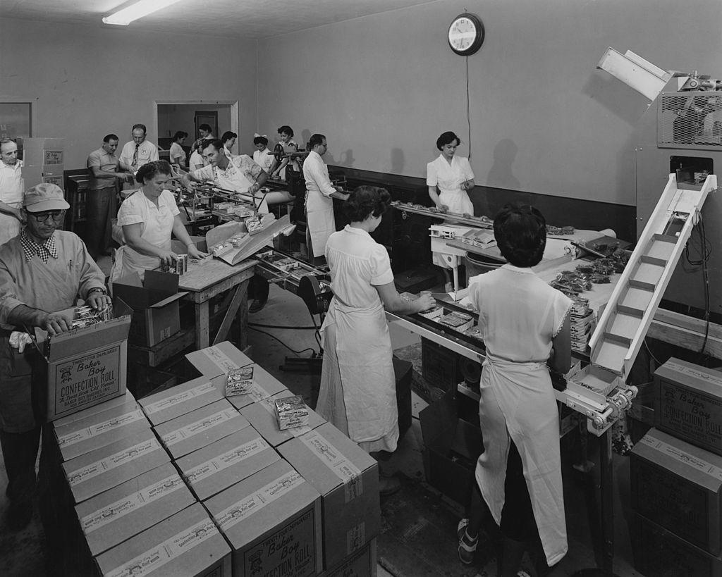 Male and female workers on a Baker Boy production line, with a man packing and sealing boxes of Baker Boy Confection Rolls to the left of the image, United States, circa 1960.