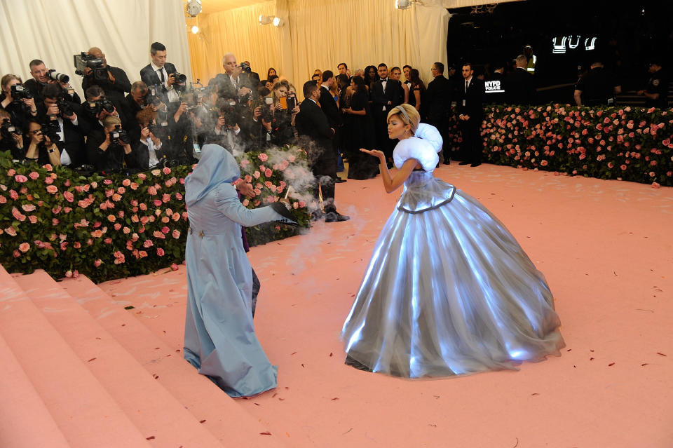 Two individuals at an event, one in a voluminous silver gown, the other in a blue outfit, with photographers in the background