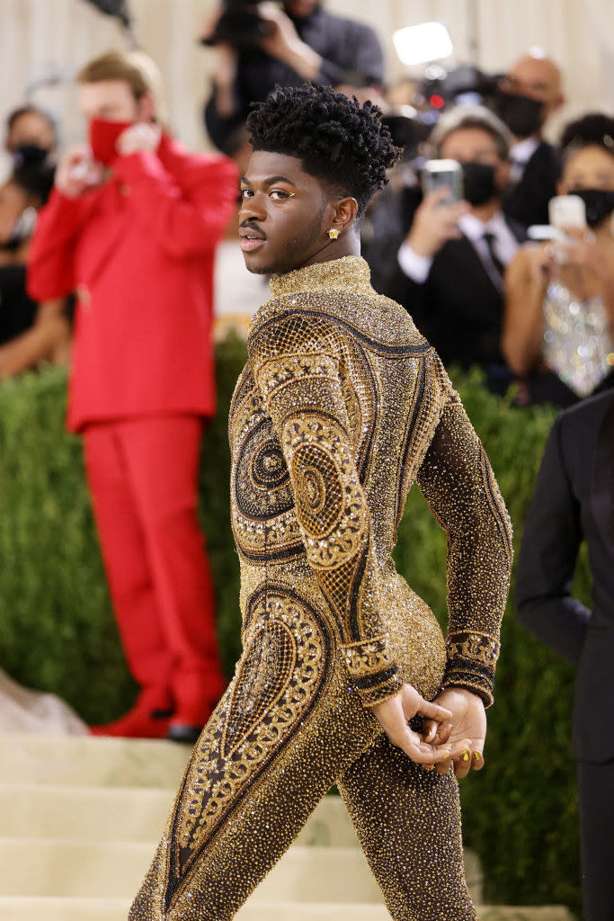 Lil Nas X poses by looking over his shoulder while wearing a long sleeve glittery jumpsuit