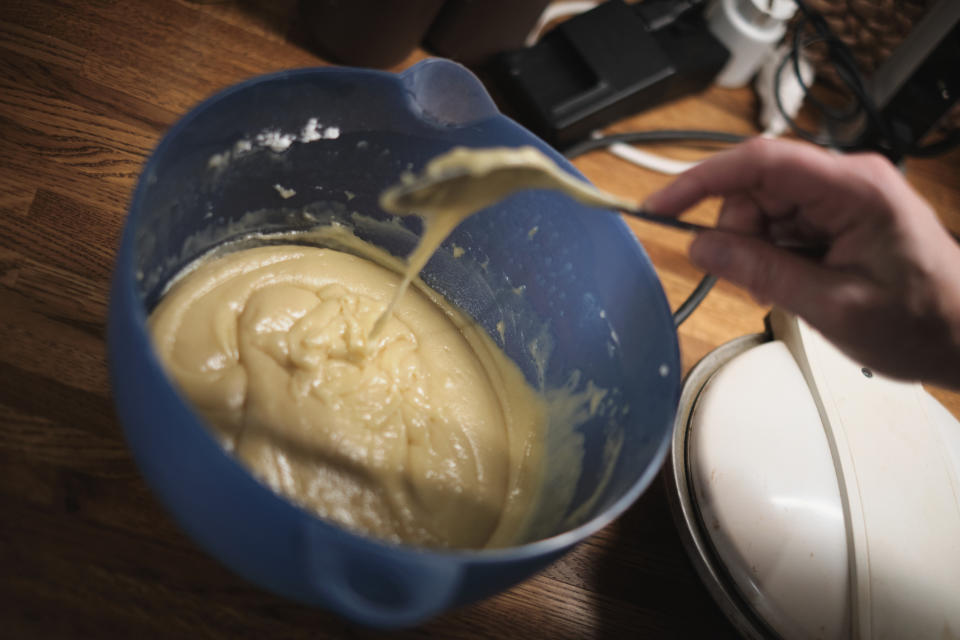 A hand holding a spoon drips batter into a mixing bowl on a wooden countertop. Various kitchen appliances are seen in the background