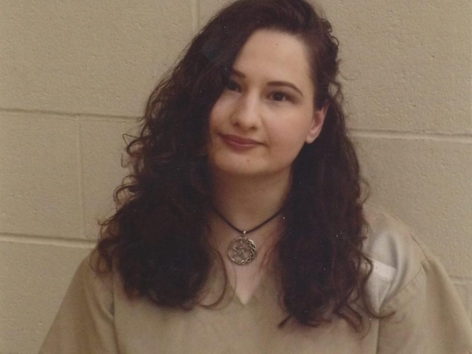 gypsy rose blanchard in prison, wearing a beige uniform and a necklace with a silver pendant. her hair is long, brown, and curly, and she's smiling