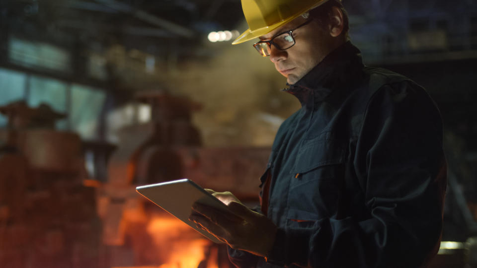 A man writing in a notebook with a steel furnace in the background.