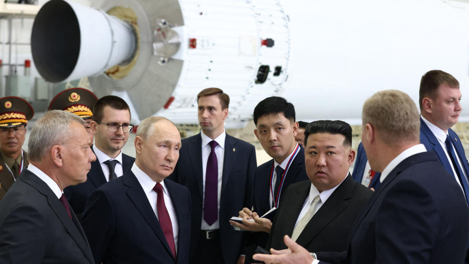 a group of men in suits talking in front of a horizontal rocket