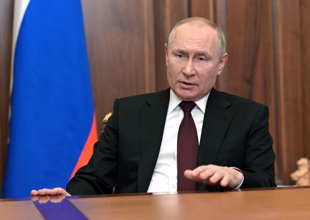 Russian President Vladimir Putin speaking while seated at a table with a flag behind him.