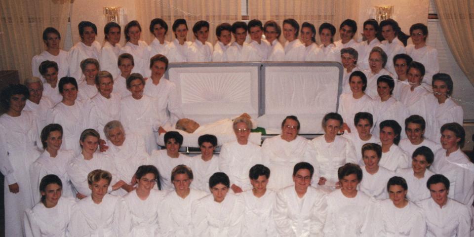 Cult leader Rolun Jeffs, self-proclaimed prophet of the FLDS, lies dead in his casket while female members of the cult gather around it and pose for a formal portrait.