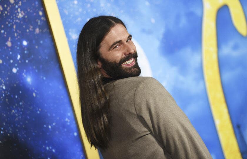 Jonathan Van Ness is smiling while looking over his shoulder and is wearing a brown jacket