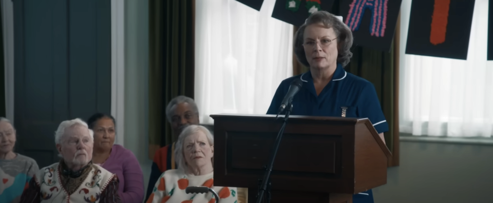 Sister Gilpin, dressed in a nurse's uniform, speaks at the podium, with the audience in the background