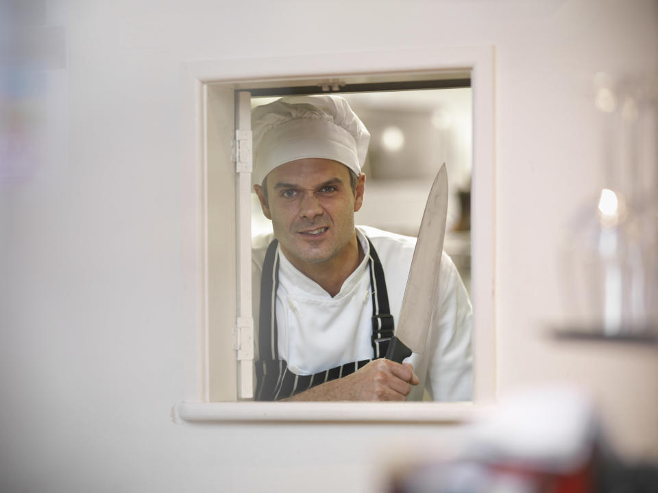 A chef in a white uniform and a hat, standing in a kitchen window, smiles while holding a large knife