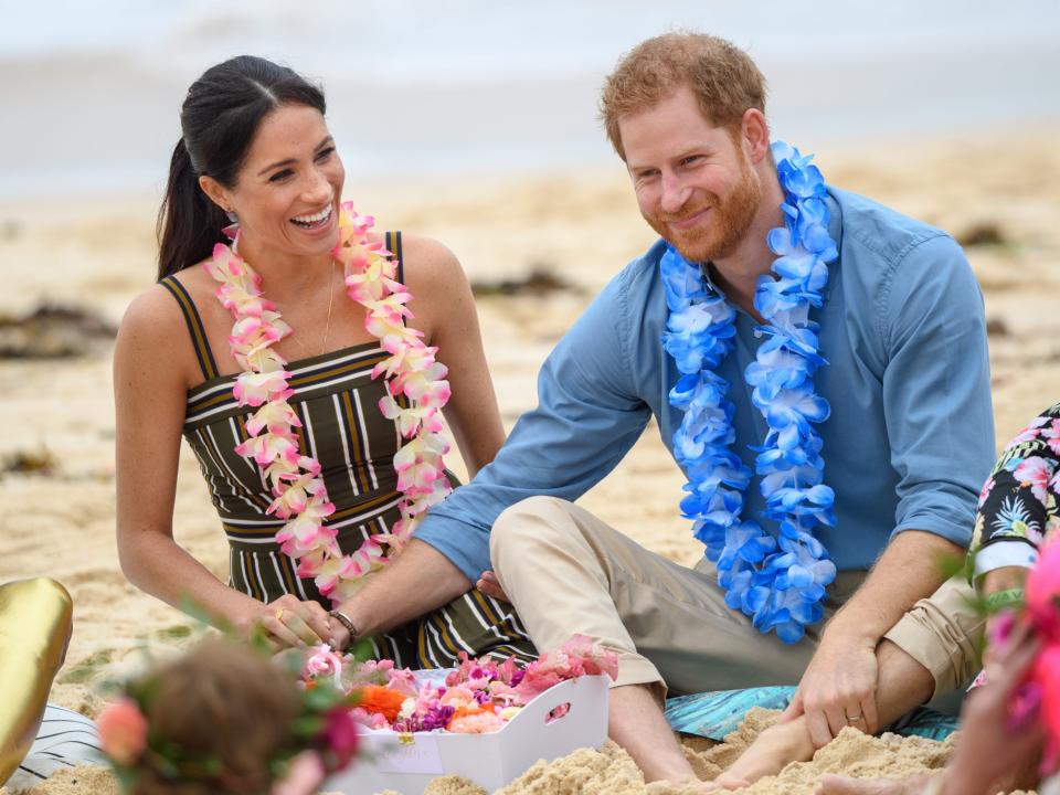 Meghan and Harry sitting on the beach with their legs crossed and holding each others hands. They're smiling and wearing casual clothing with flower necklaces.