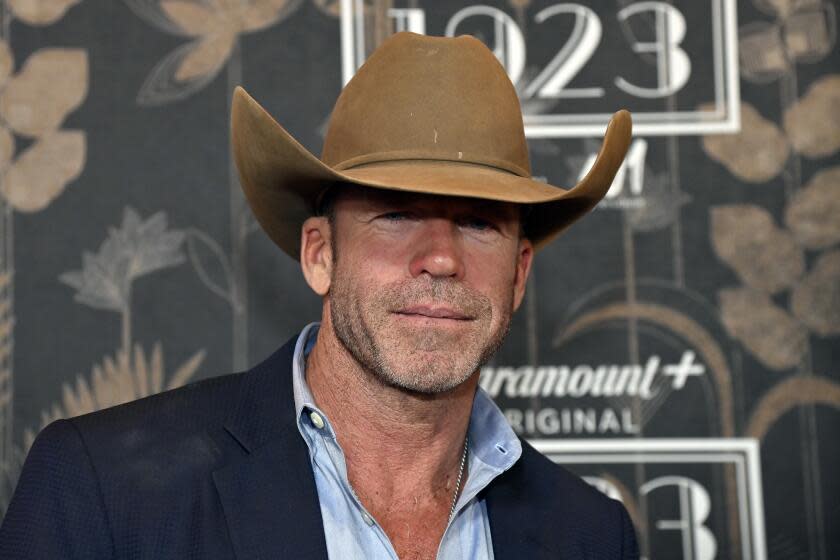 Taylor Sheridan smiles in a blue coat and shirt as a light brown cowboy hat casts a shadow on his eyes