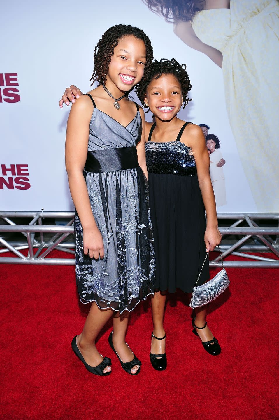 chloe bailey and halle bailey smile at the camera while standing on a red carpet, the young girls wear sleeveless formal dresses and black shoes, halle holds a purse