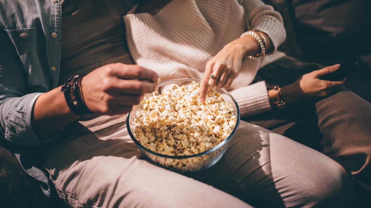 Close-up of chic couple's hands watching television and eating popcorn at night.