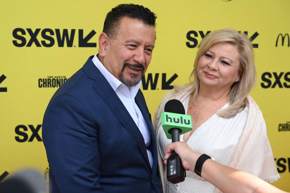 richard montañez, wearing a blue suit and white shirt, smiling and speaking into a microphone while standing next to judy montañez, who wears a white dress