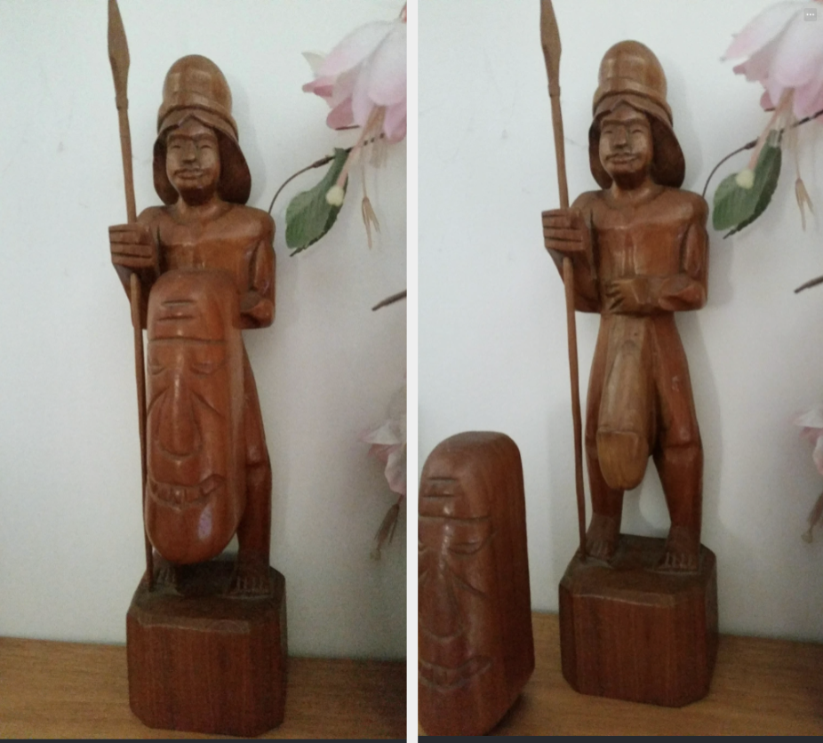 A wooden sculpture depicts an indigenous warrior with a spear, wearing a helmet, and holding a shield. The sculpture is decorated with flowers in the background