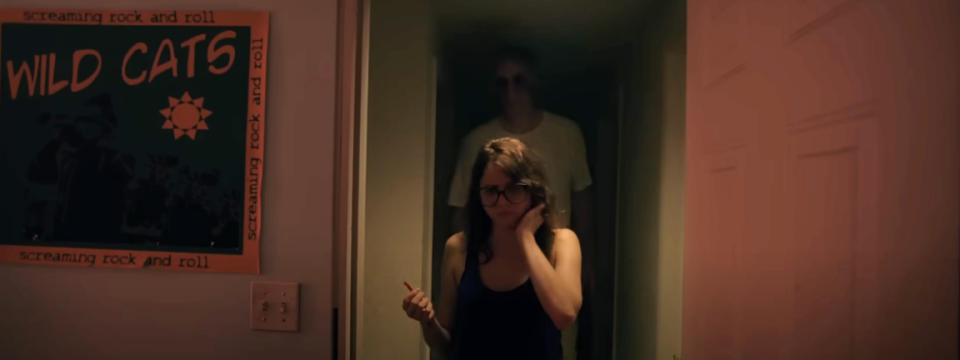 A scene from a movie shows a concerned woman walking through a dimly lit hallway with a shadowy figure in the background. A poster for "Wild Cats" is visible on the wall