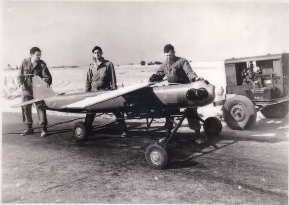 In a black and white photo, three men look at what appears to be a very small unmanned plane.