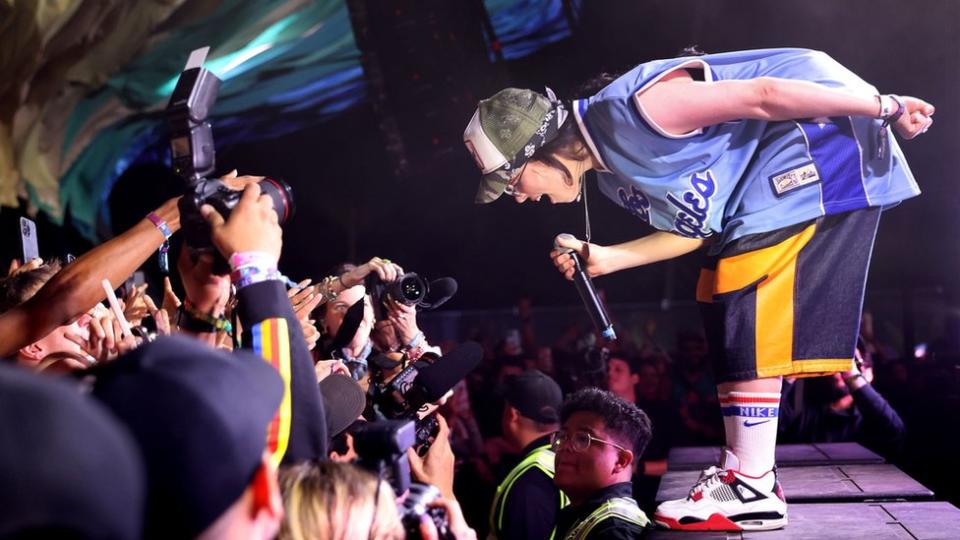 Billie Eilish performs at the Coachella festival.  Billie is a 22-year-old white woman with shoulder-length black hair.  She is depicted on stage, leaning towards the crowd.  She wears blue and yellow sports shorts, matched with a blue sports jersey, white sports socks and white sneakers with red trim.  She wears a green and white cap over a blue bandana and holds the microphone in her right hand while her left is behind her back.  A line of security guards separates her from the otherwise very close crowd.  People in the crowd hold up their phones and cameras to capture his performance.
