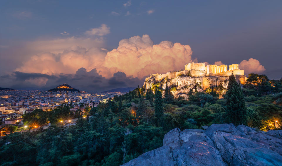 The image shows a scenic view of the Acropolis in Athens at dusk, with city lights and lush greenery in the foreground, and dramatic clouds in the sky