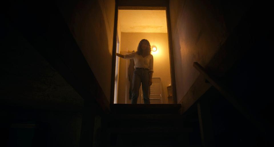 A young woman stands before a dark basement staircase in an illuminated doorway