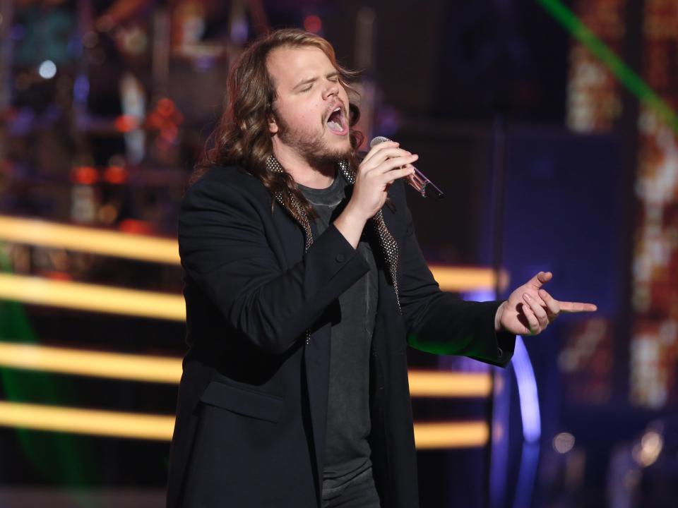 Caleb Johnson performs onstage in a black suit with yellow lights in background