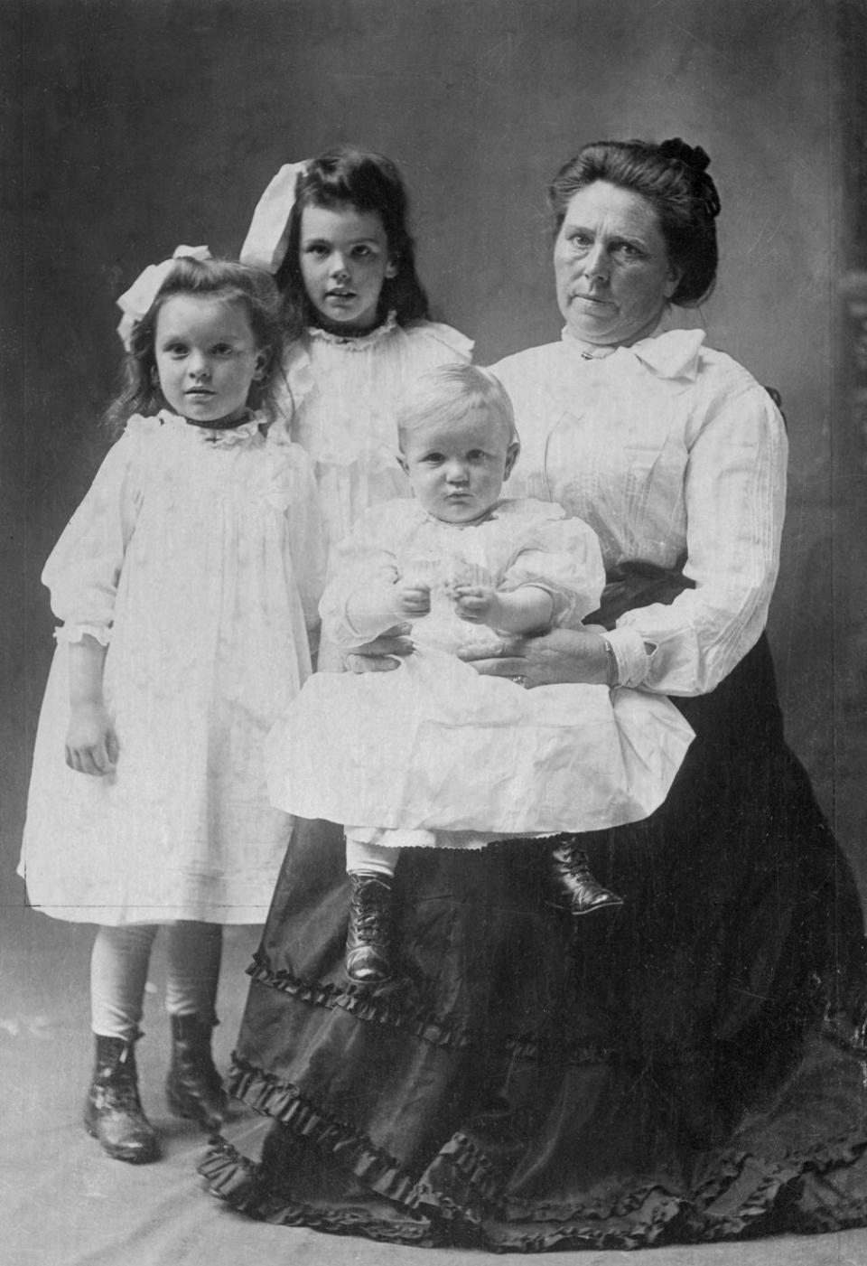 two young girls stand next to belle gunness who is seated and holds a baby on her lap, all three children wear white dresses, gunness wears a long sleeve light colored blouse and a dark floor length skirt