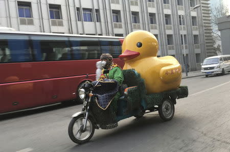 A man wearing a mask and dressed in a clown costume rides an electric tricycle while carrying a container in the shape of a rubber duck amid heavy smog in Beijing, China, December 29, 2015. REUTERS/Jason Lee