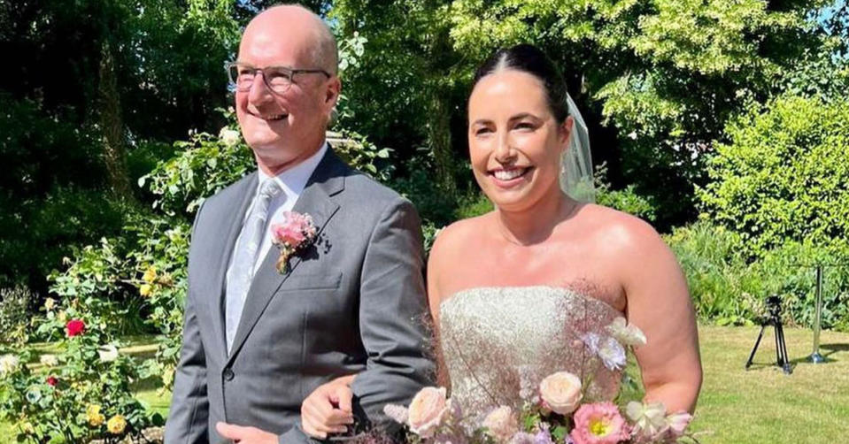 Sunrise host Kochie walks his daughter Georgie down the aisle. He is wearing a grey suit with a pink flower. She is wearing a strapless wedding dress.