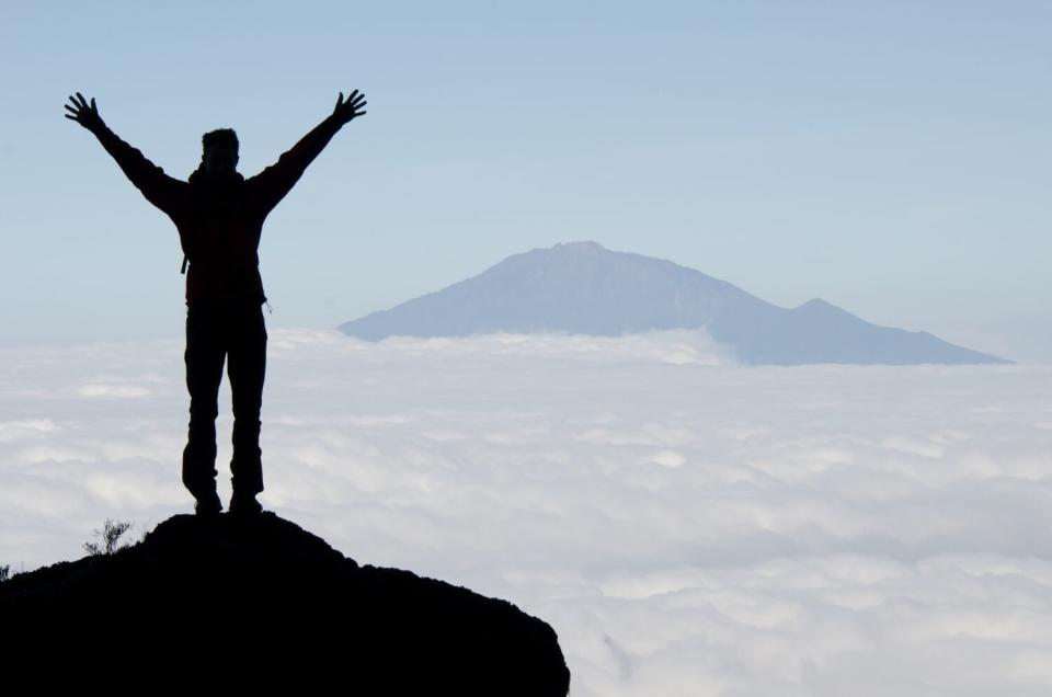 A person, seen in silhouette, stands with arms outstretched amid clouds while facing a mountain peak in the distance