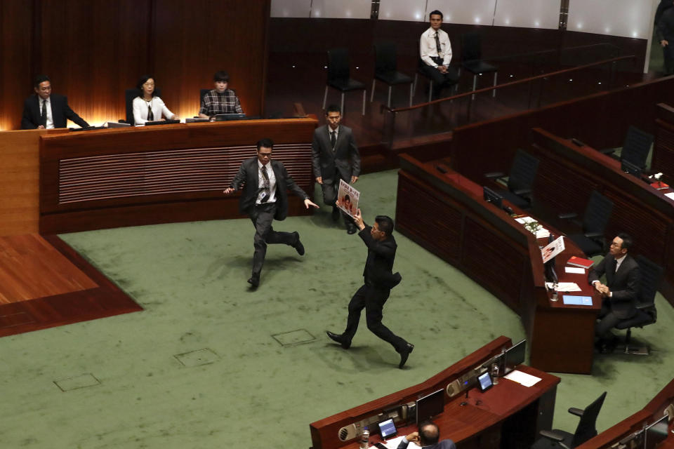 Security officials run to stop a pro-democracy lawmaker holding a plate card with picture of Hong Kong Chief Executive Carrie Lam with bloody hand on her face and slogan " Five Demands, No One Less" approach toward to Carrie Lam during a question and answer session with lawmakers at the chamber of the Legislative Council in Hong Kong, Thursday, Oct. 17, 2019. (AP Photo/Mark Schiefelbein)