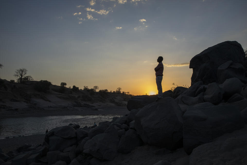 Surgeon and doctor-turned-refugee, Dr. Tewodros Tefera, stands at the Tezeke River crossing in Hamdayet, Sudan, on March 16, 2021. At left across the river is Ethiopia. He plans to continue his work in Sudan. The high-rise buildings of his city in Tigray, Humera, can be seen on the horizon. He could walk home, but he’s not sure he will ever go there again. (AP Photo/Nariman El-Mofty)