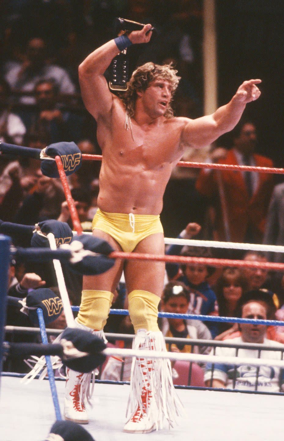 kerry von erich stands inside a wrestling ring holding a belt over his head with one hand and pointing right with his other, he wears yellow shorts and knee pads with knee high white boots with long fringe and red laces