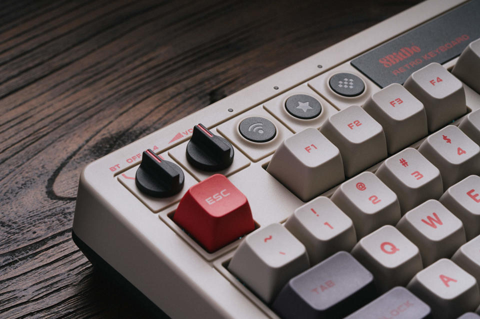 Produktová fotografia mechanickej klávesnice 8BitDo Retro.  Detailný záber na klávesnicu vľavo hore.  Má čierne spínače a šedobiele, červené alebo tmavosivé klávesy.