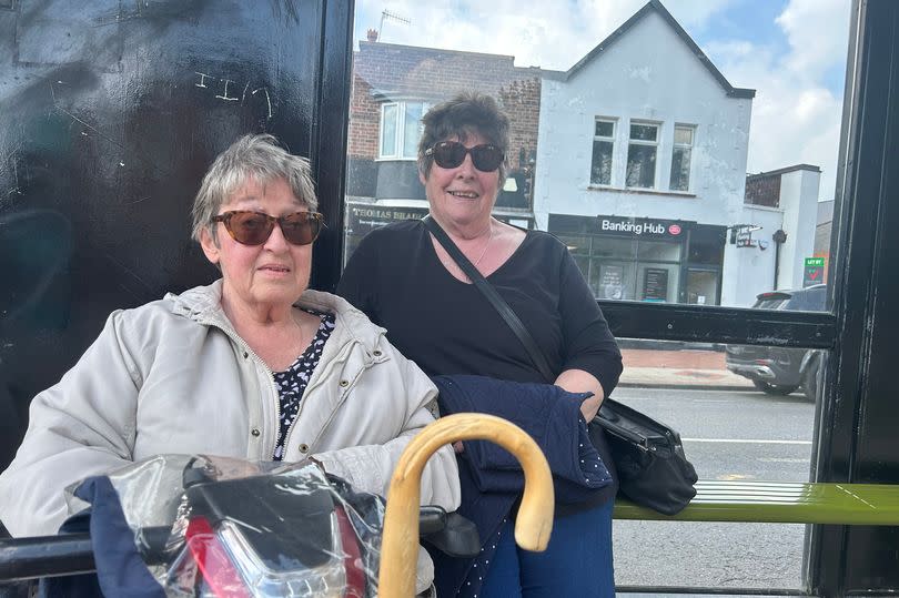 Paula Gibson (L) on mobility scooter and Margaret Williamson (R) sitting, smiling at bus stop outside Post Office Banking Hub on Derby Road, Stapleford