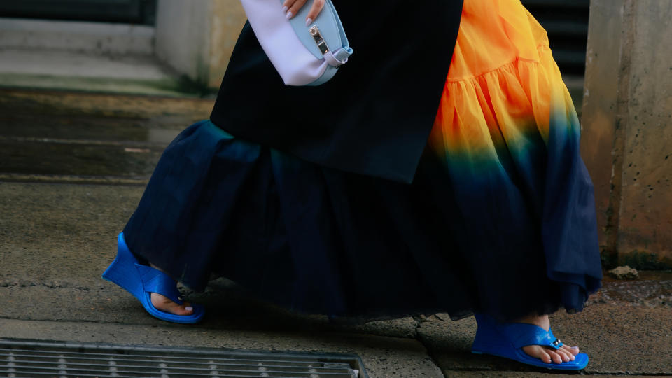 Violet Grace wearing black coat, white oversized floral broach, black skinny sunglasses, Ferragamo pale blue clutch, orange and blue tie dyed skirt and blue heels Australian Fashion Week Presented By Pandora 2024 at Carriageworks on May 13, 2024 in Sydney, Australia.