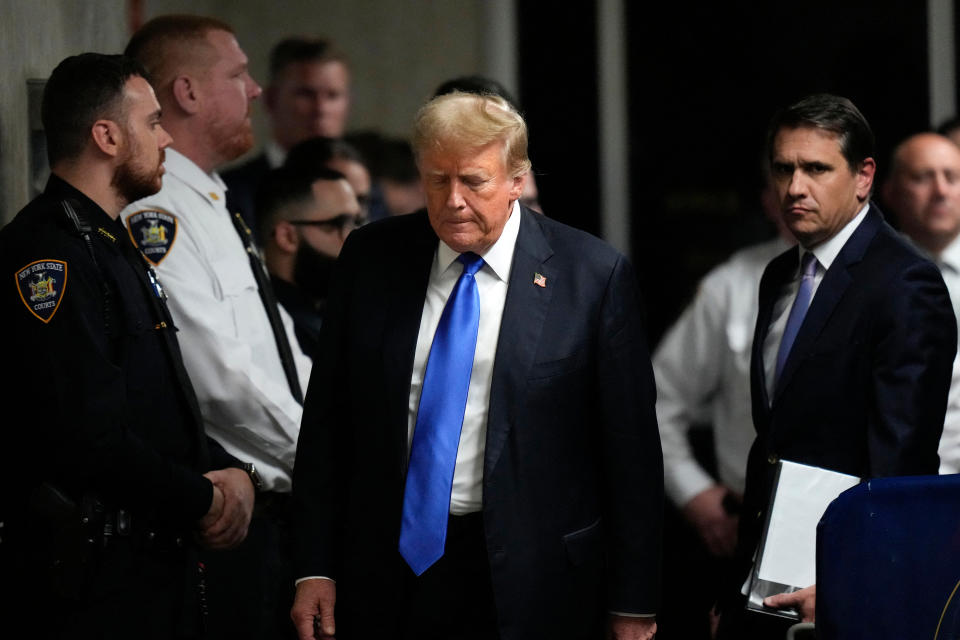 Donald Trump is surrounded by police officers and security personnel indoors, appearing deep in thought