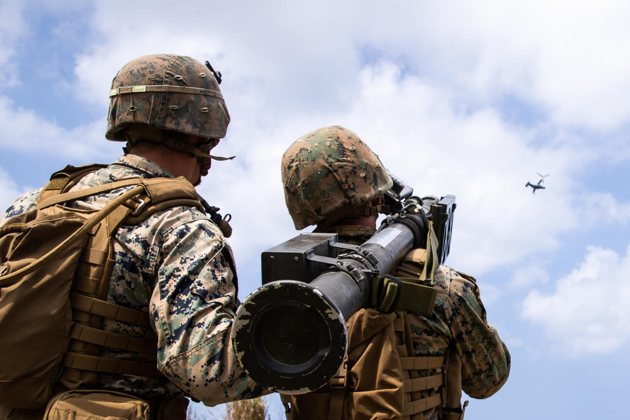 Two service members, from behind, wear camouflaged helmets and fatigues. One holds a shoulder-mounted surface-to-air missile as they face toward a lone plane against white clouds in a blue sky.
