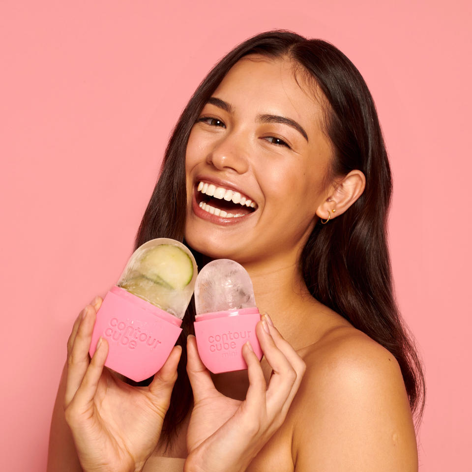 Against a pink background, an olive skinned brunette woman smiles while holding two pink moulds of ice. 