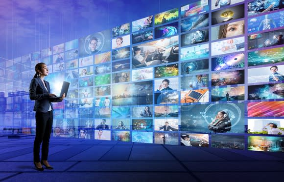 Woman holding a PC looks at a wall of TV screens.