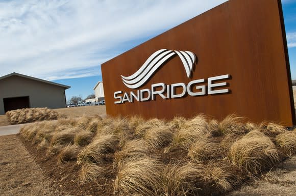 SandRidge logo on a wood-colored sign, with brush and industrial buildings in the background.