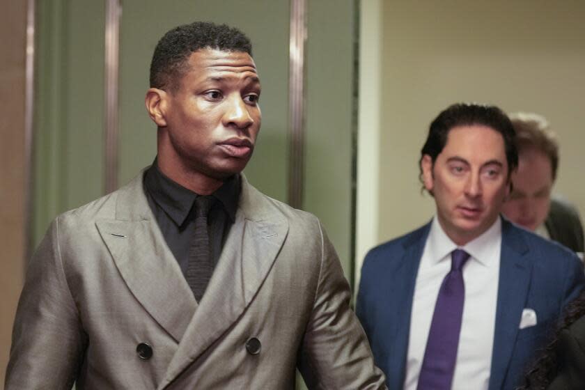 Jonathan Majors standing in front of a door in a grey suit looking to his left in front of a man in a blue suit jacket