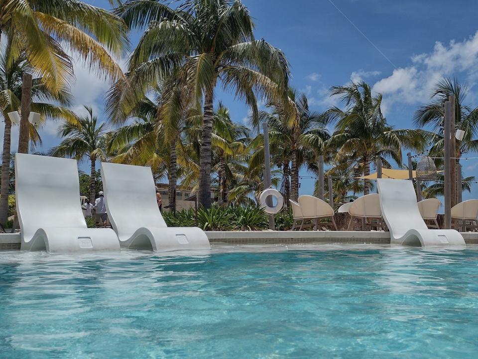 Shot of a pool with three white loungers backdropped by palm trees