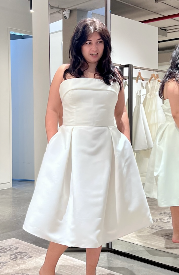 Person in a bridal store trying on a strapless, knee-length white dress with a full skirt