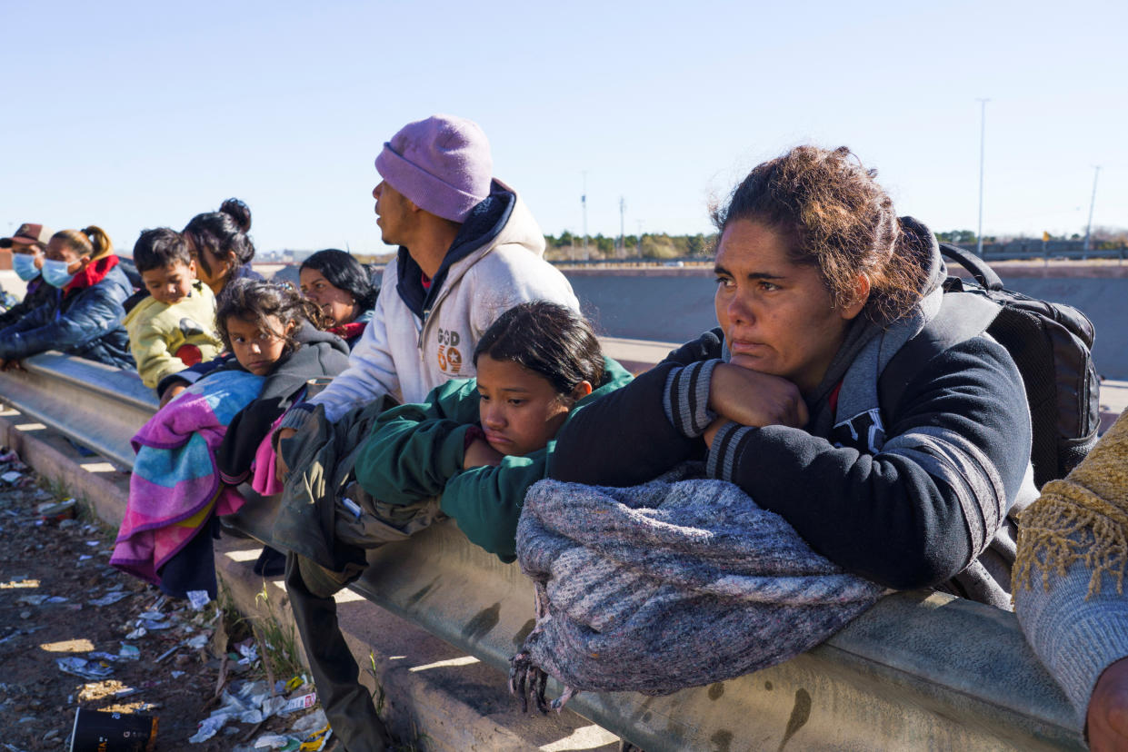 Migrants stand in line along a metal barricade.