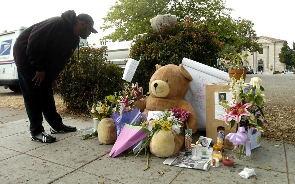 A person looks at a street memorial of flowers, notes and a teddy bear.