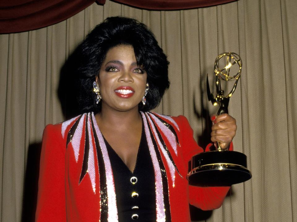 Oprah in a red and black coat and dress holding an Emmy award.