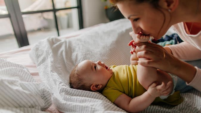 Portrait of a little smiling baby boy and his mom, kissing his tiny feet right after waking up in his nursery.