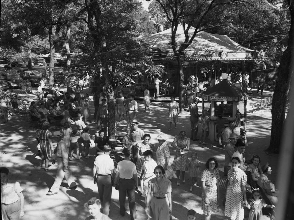 August 1941: With the summer season winding down, the Forest Park Zoo and amusement park remain one the most popular attractions in Fort Worth. Pictured is the view across the amusement grounds. Adults are enjoying the shade from the trees as they wait for their children on the rides. There is a ticket counter in the middle of the concourse and a Merry-go-round in the background. The parking lot is in the back left of the photograph.