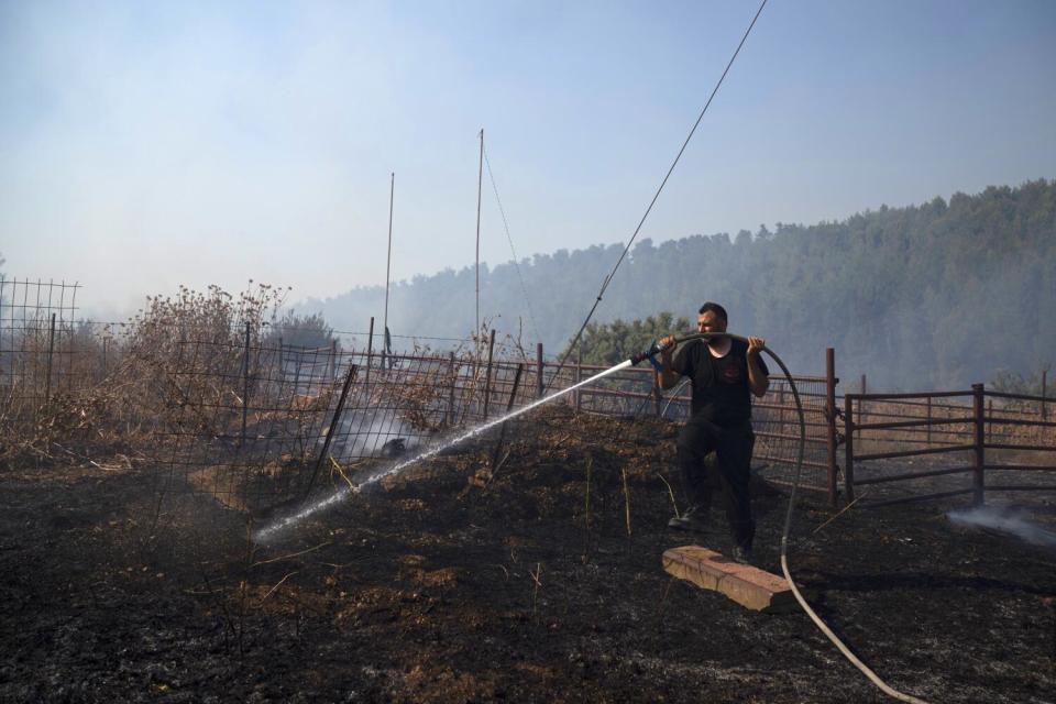 A man sprays water form a hose onto smoldering ground near a fence.