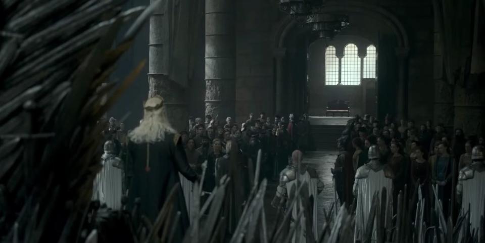 Viserys stands in front of the throne in a crowded THrone Room and looks towawrds the open doors
