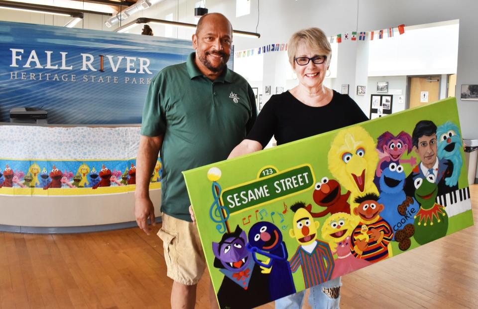 Artist Sheila Oliveira and Visitor Services Supervisor at Fall River Heritage State Park Jim Lopes stand in the visitors center lobby with Oliveira's portrait of Fall River composer Joe Raposo, who wrote the theme song for Sesame Street.